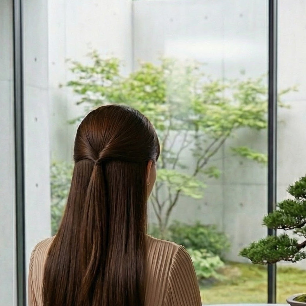 Kewevia Owner. Woman in a beige dress sitting at a table looking out at a bonsai tree through large windows.